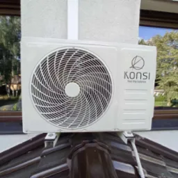 Zewnętrzna jednostka klimatyzacji marki KONSI zamontowana na wspornikach na dachu domu, widoczne okno z odbijającym się drzewem.