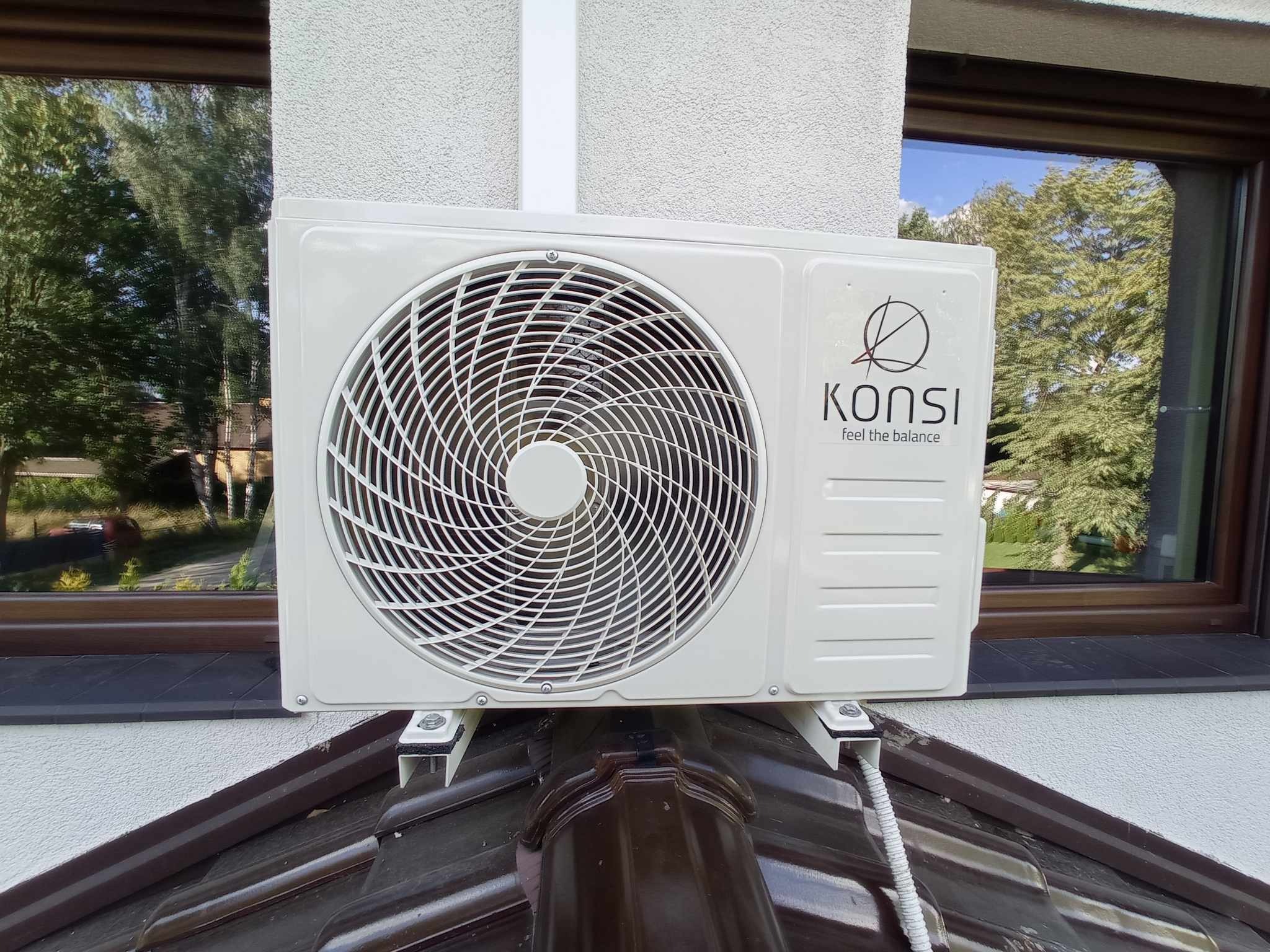 Zewnętrzna jednostka klimatyzacji marki KONSI zamontowana na wspornikach na dachu domu, widoczne okno z odbijającym się drzewem.