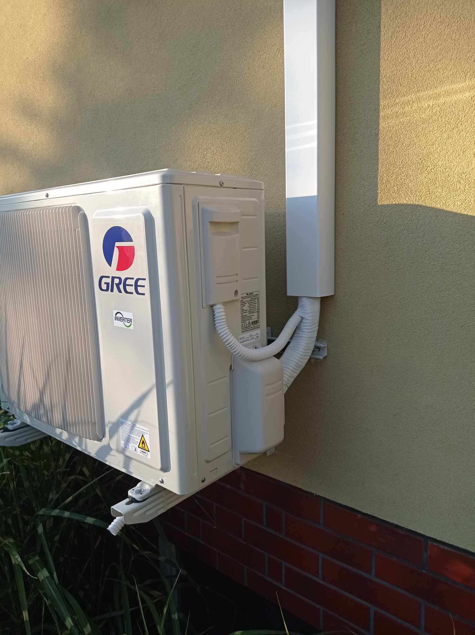 Zewnętrzna jednostka klimatyzacji marki Gree zamontowana na elewacji budynku z ceglaną podmurówką, widoczne rury odprowadzające skropliny i peszle z przewodami.