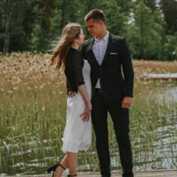Para młoda na drewnianym pomoście nad jeziorem, panna młoda w białej sukience z ażurowym wzorem i czarnym bolerku, pan młody w czarnym garniturze z wzorzystą koszulą, w tle trzciny i drzewa.
