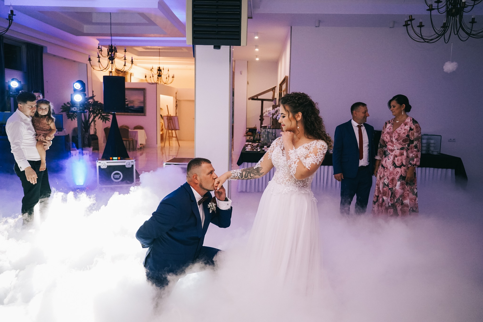 Pan młody klęczy i całuje rękę panny młodej podczas pierwszego tańca w chmurach, goście obserwują, dekoracje weselne w tle.