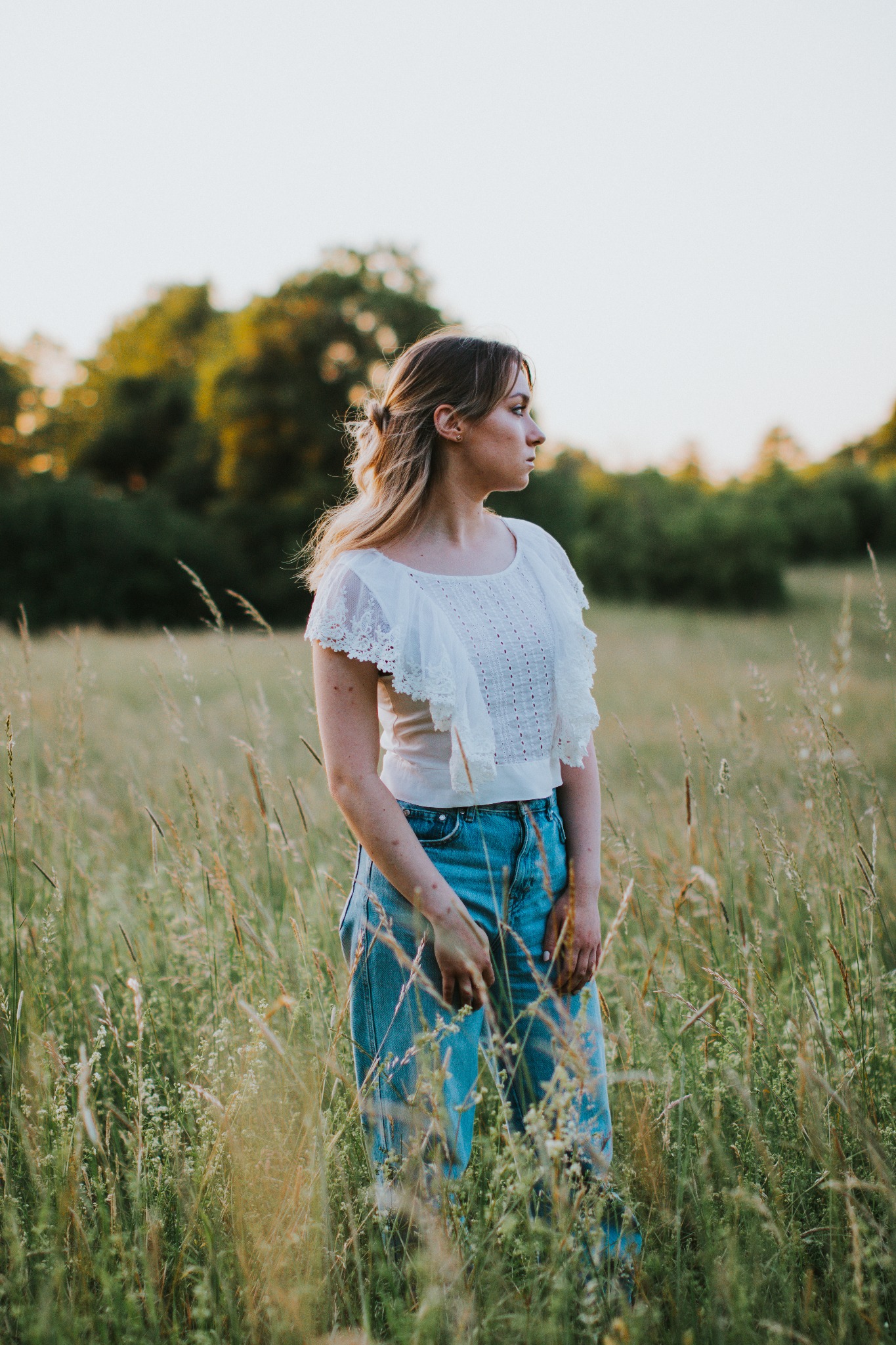 Portret kobiety w białej bluzce z koronkowymi falbanami i dżinsach, stojącej na łące w promieniach zachodzącego słońca, z profilu w lewo.