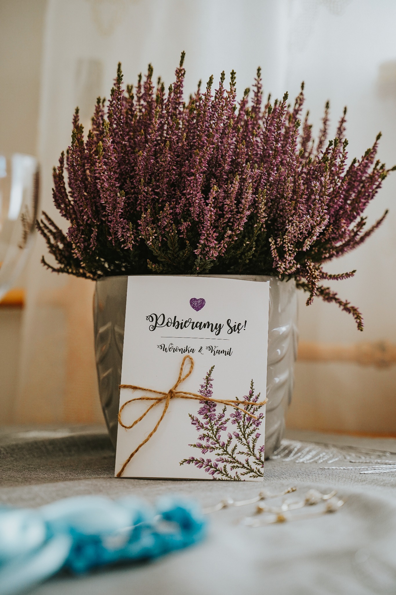 Zaproszenie ślubne z motywem wrzosu, przewiązane sznurkiem, ustawione przed doniczką z wrzosem na szarym obrusie.