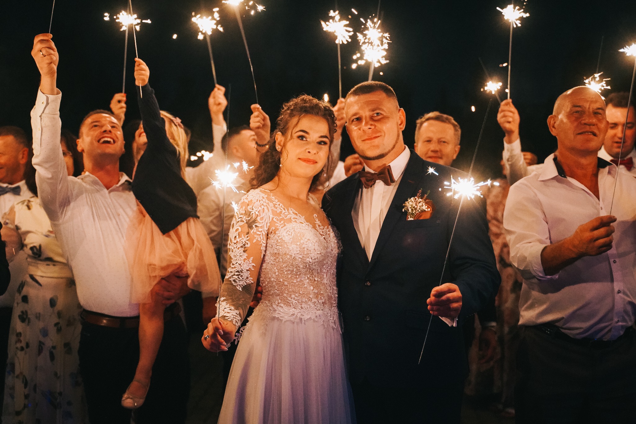 Para młoda pozuje z gośćmi weselnymi trzymającymi zapalone zimne ognie, ciemne tło, wieczorowa atmosfera.
