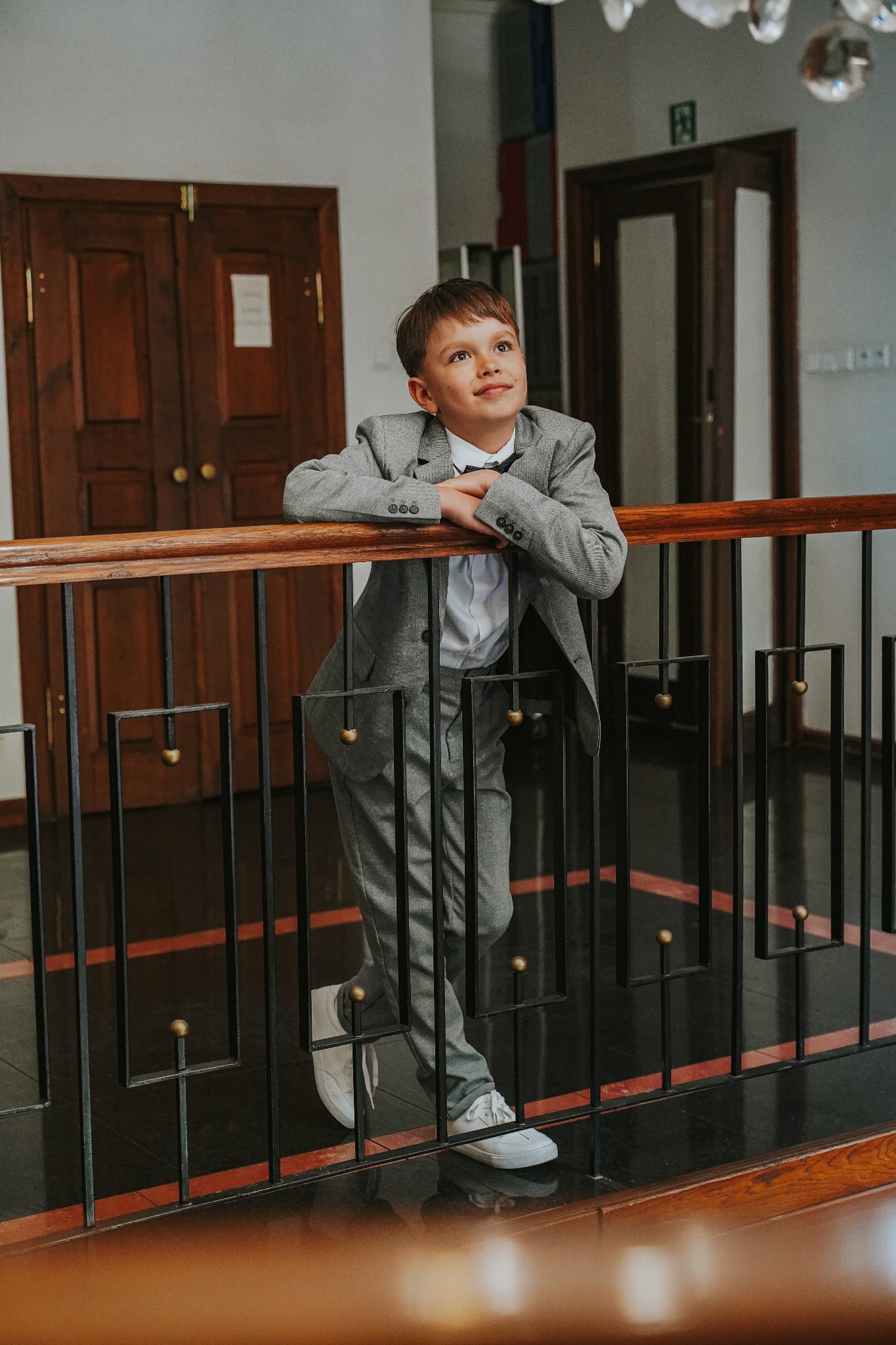 Chłopiec w szarym garniturze i białych sneakersach opiera się o drewnianą balustradę, spoglądając w górę w jasnym wnętrzu z ciemnymi drewnianymi drzwiami w tle.