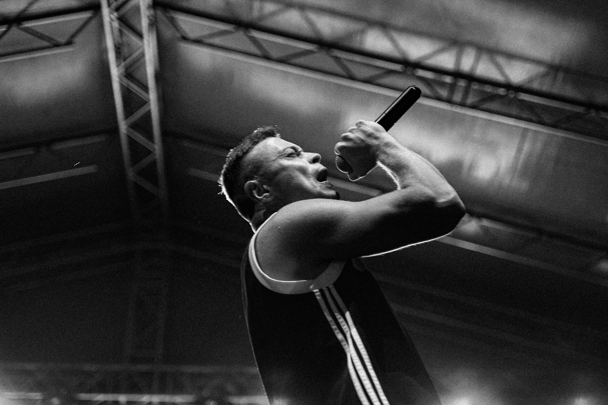 Czarno-białe zdjęcie piosenkarza z mikrofonem na scenie, skierowanego w górę, z widoczną konstrukcją dachu. Artysta z zaciśniętą dłonią trzyma mikrofon, z otwartymi ustami w trakcie występu.