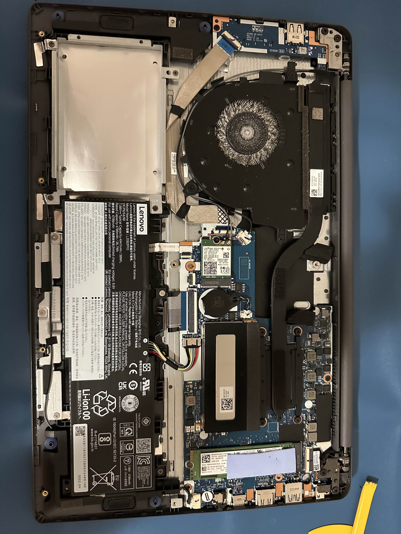 Wnętrze otwartego laptopa Lenovo z widocznymi komponentami: baterią, wentylatorem, dyskiem, płytą główną i przewodami, na niebieskim tle.