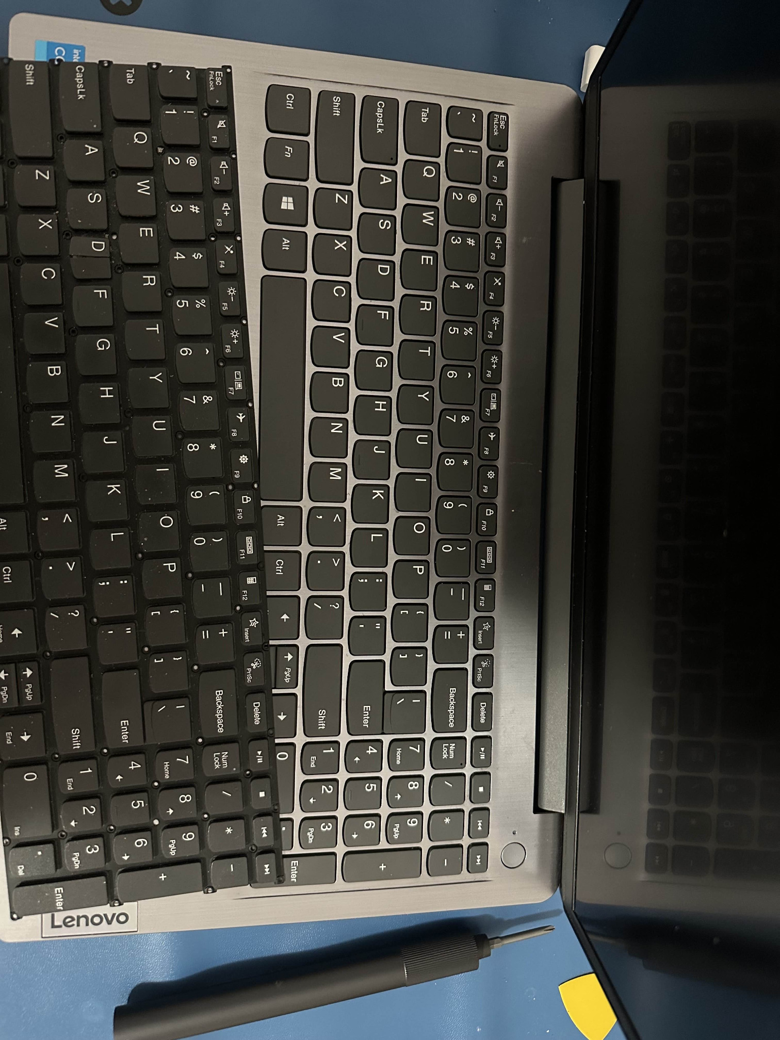 Laptop Lenovo z widoczną, częściowo zdemontowaną klawiaturą, obok leży śrubokręt precyzyjny i plastikowy otwierak.