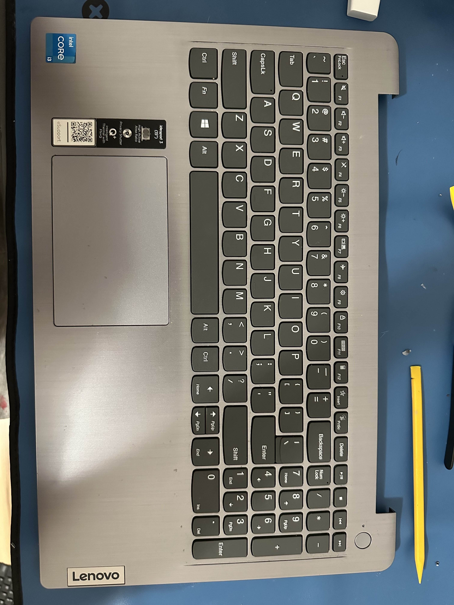 Widok z góry na częściowo zdemontowany laptop Lenovo Ideapad 3 z widoczną klawiaturą, touchpadem, naklejkami informacyjnymi (Intel Core i3, eSupport), obok leżą narzędzia do demontażu.