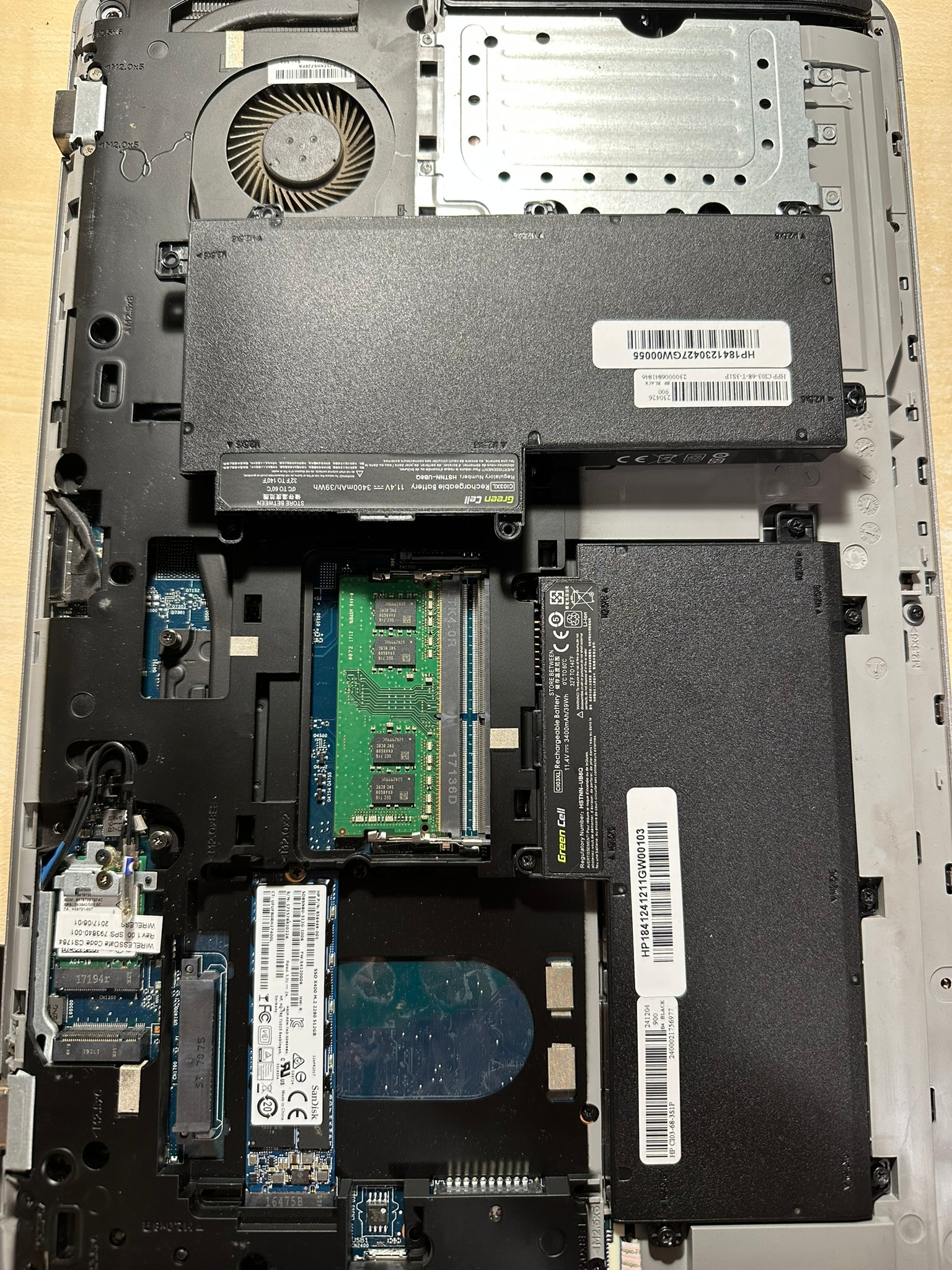 Wnętrze laptopa HP z widocznymi komponentami: baterią, wentylatorem, modułami pamięci RAM i dyskiem SSD. Widoczne oznaczenia i numery seryjne podzespołów.