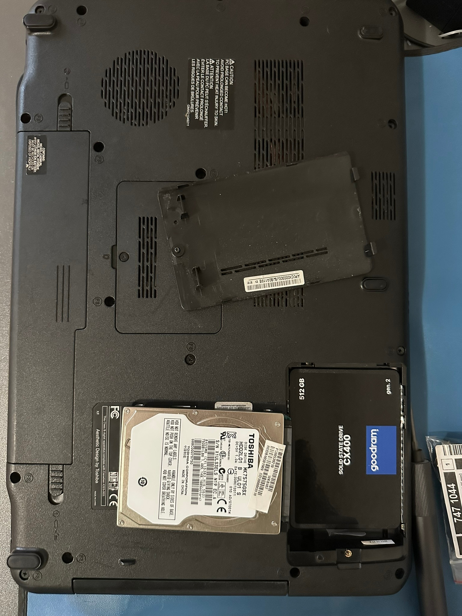 Otwarty laptop z widocznym dyskiem HDD Toshiba i dyskiem SSD Goodram CX400 512GB. Widoczna pokrywa dysku. Detaliczny widok wnętrza komputera.