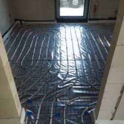 Instalacja ogrzewania podłogowego w trakcie montażu, rury grzewcze ułożone na folii izolacyjnej z siatką, widoczne mocowania i okno dachowe z odbijającym się samochodem.