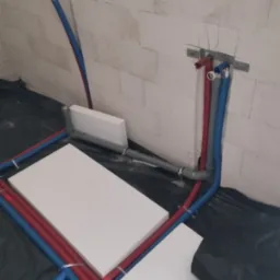 Instalacja wodno-kanalizacyjna w trakcie montażu: rury czerwone i niebieskie prowadzone po izolacji podłogowej, widoczne uchwyty mocujące i fragment ściany z białych bloczków.