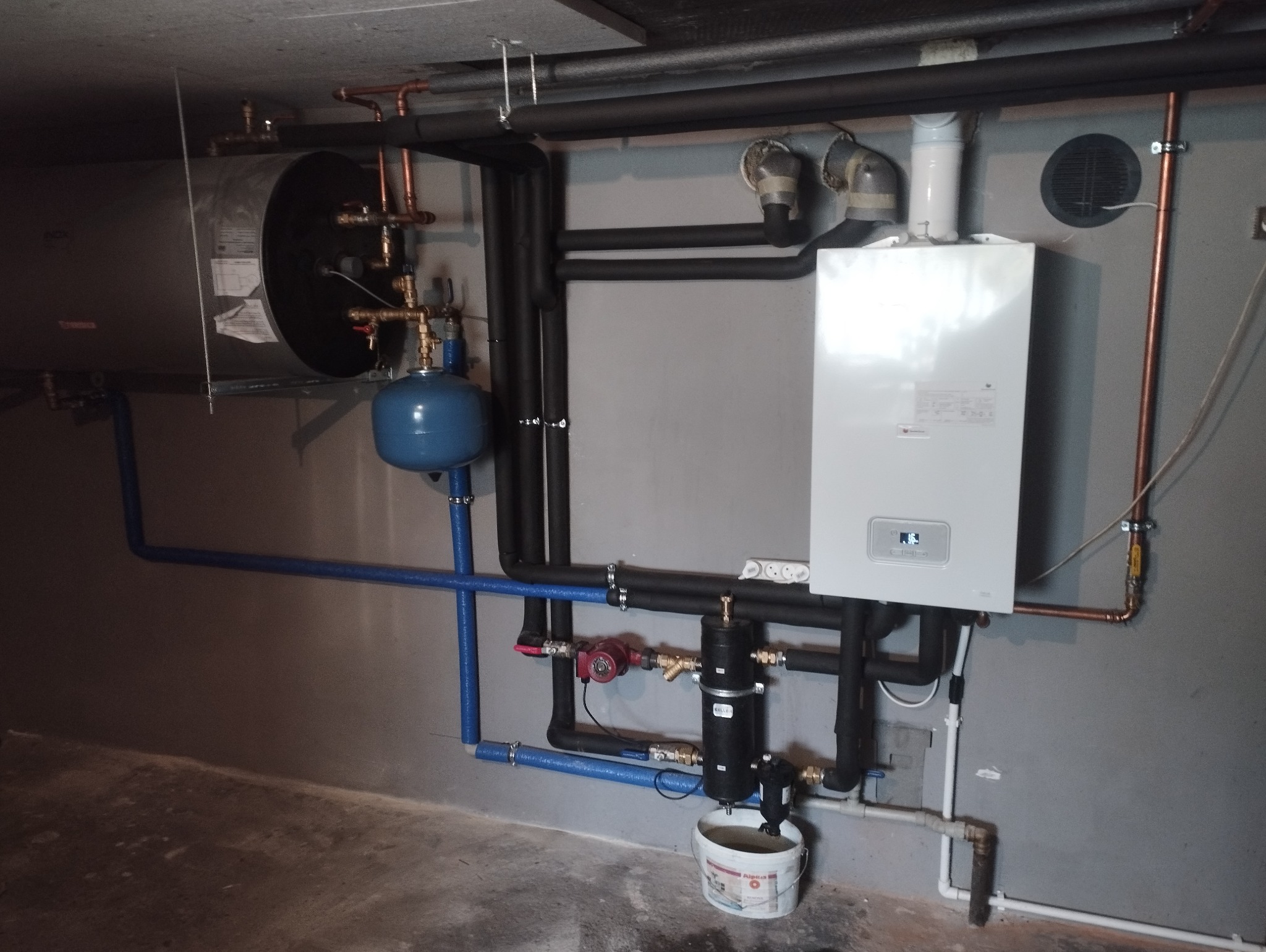 Zakończona instalacja grzewcza z białym piecem gazowym, zbiornikiem akumulacyjnym, niebieskimi rurami i czarnymi rurami izolowanymi, widoczne elementy instalacji hydraulicznej, filtr i pompa, w tle...