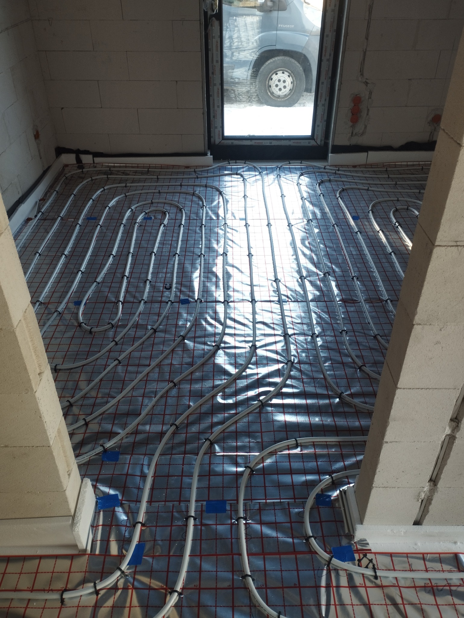 Instalacja ogrzewania podłogowego w trakcie montażu, rury grzewcze ułożone na folii izolacyjnej z siatką, widoczne mocowania i okno dachowe z odbijającym się samochodem.
