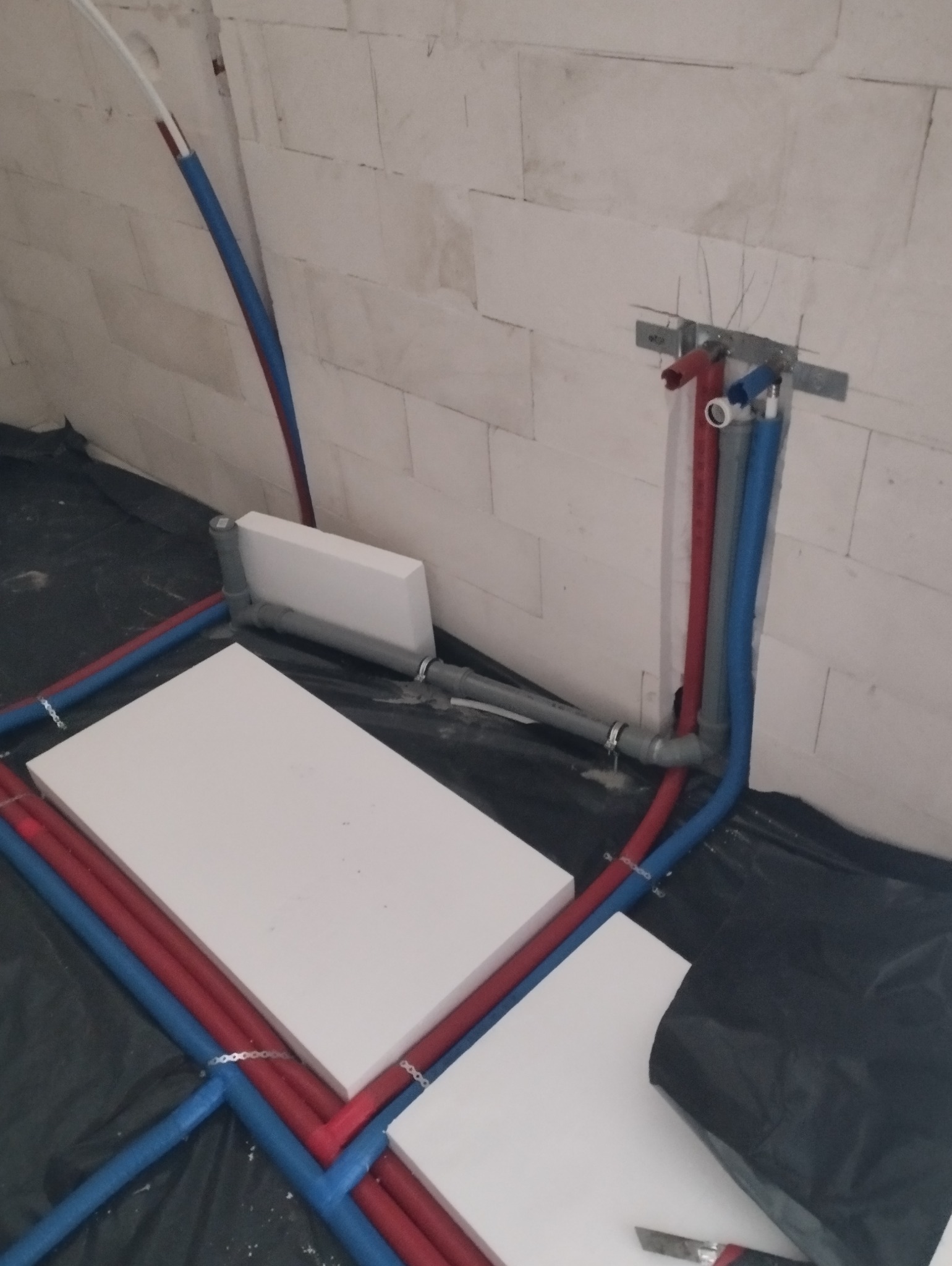 Instalacja wodno-kanalizacyjna w trakcie montażu: rury czerwone i niebieskie prowadzone po izolacji podłogowej, widoczne uchwyty mocujące i fragment ściany z białych bloczków.