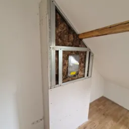 Ocieplenie poddasza wełną mineralną i zabudowa z płyty karton-gips z widocznymi profilami metalowymi, ukrywająca licznik gazu, na tle skośnej ściany i drewnianej belki stropowej.