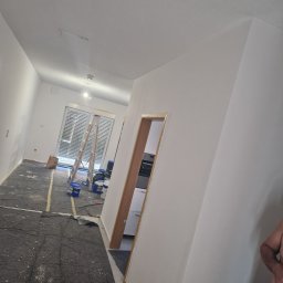 Bau Servis - Wnętrze mieszkania w trakcie malowania ścian na biało, widoczna drabina oparta o okno, zakryta podłoga, puszki z farbą i fragment sylwetki malarza.