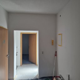 Bau Servis - Wnętrze w trakcie remontu: świeżo pomalowane ściany na szaro, zabezpieczone taśmą framugi drzwi, widoczna szpachla i narzędzia malarskie przy ścianie.