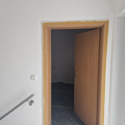Bau Servis - Przygotowanie do malowania ścian i futryny drzwiowej z użyciem taśmy malarskiej w Katowicach. Widoczny fragment schodów z metalową barierką oraz zabezpieczona podłoga w pomieszczeniu za drzwiami.