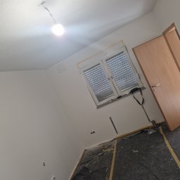 Bau Servis - Pokój w trakcie remontu: ściany przygotowane do malowania, okno zabezpieczone taśmą malarską, podłoga zakryta folią ochronną, widoczna wisząca lampa.