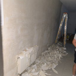 Bau Servis - Ściana po usunięciu starej powłoki, z widocznym grzejnikiem częściowo pokrytym resztkami, drabina oparta o ścianę, przygotowanie do dalszych prac wykończeniowych.