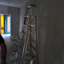 Bau Servis - Wnętrze w trakcie remontu: aluminiowa drabina oparta o ścianę, na podłodze fragmenty starej tapety, w tle widoczne drzwi z pomarańczową szybą.