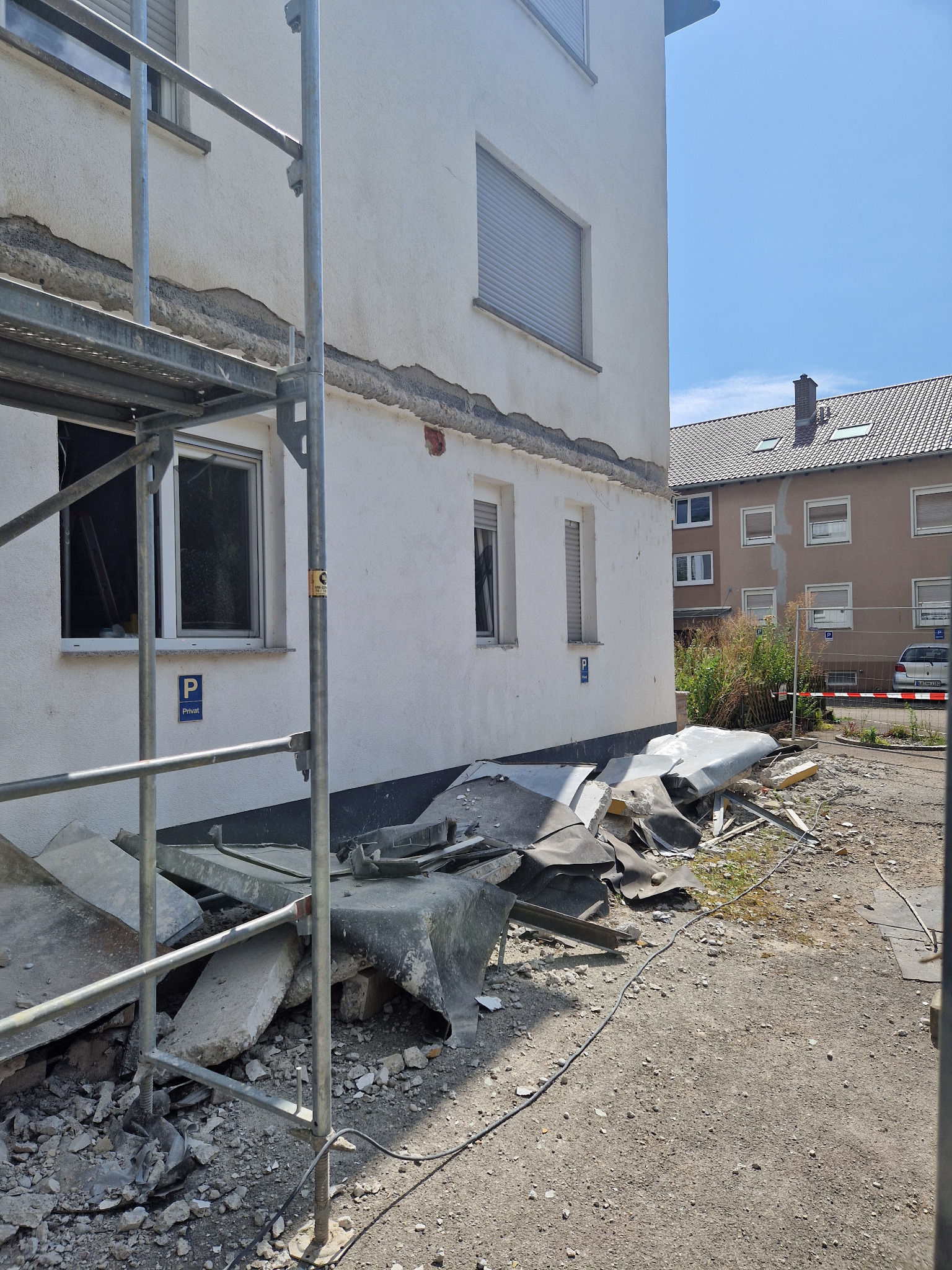 Fragment elewacji budynku w trakcie remontu, widoczne rusztowanie, uszkodzona warstwa elewacyjna i gruz na ziemi.