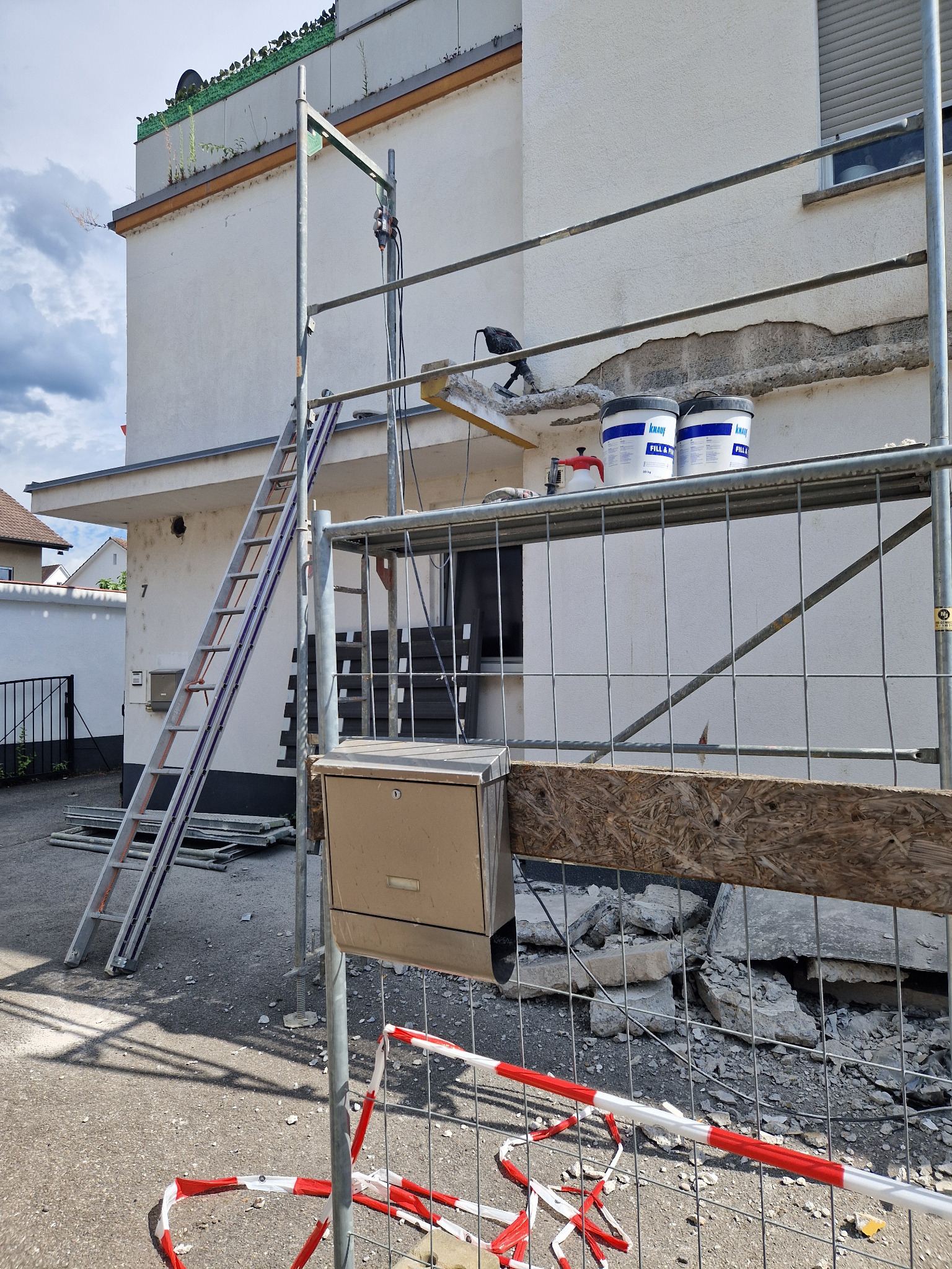 Fragment elewacji budynku w trakcie naprawy, widoczne rusztowanie, drabina, dwa wiadra z masą szpachlową, narzędzie do nakładania masy oraz odspojona warstwa tynku.