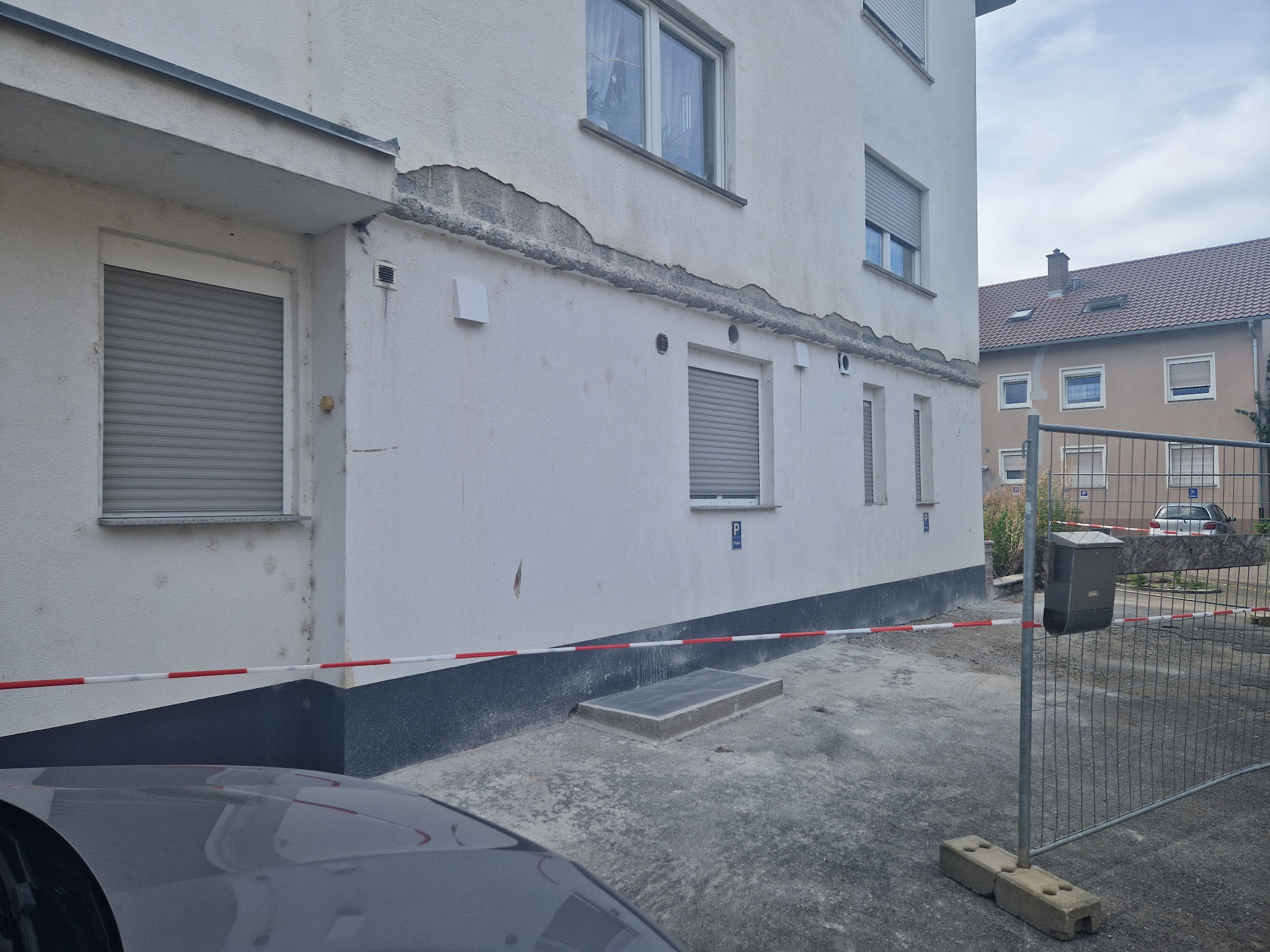 Fragment budynku z uszkodzonym tynkiem i widocznymi śladami naprawy, zabezpieczony taśmą ostrzegawczą i ogrodzeniem tymczasowym, z fragmentem samochodu na pierwszym planie.
