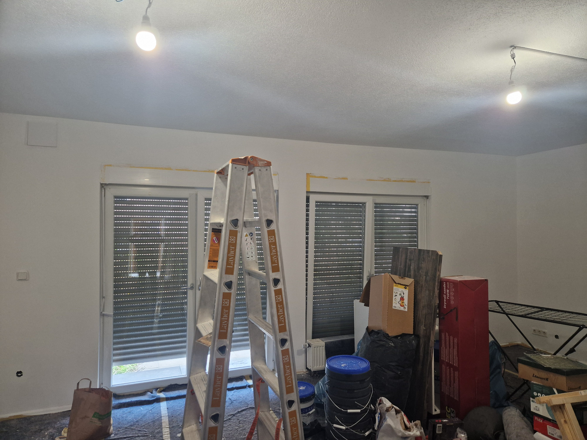 Pokój w trakcie przygotowań do prac wykończeniowych; zabezpieczone taśmą malarską framugi okienne, rozstawiona aluminiowa drabina Layher, podłoga zabezpieczona folią, widoczne puszki z farbą...