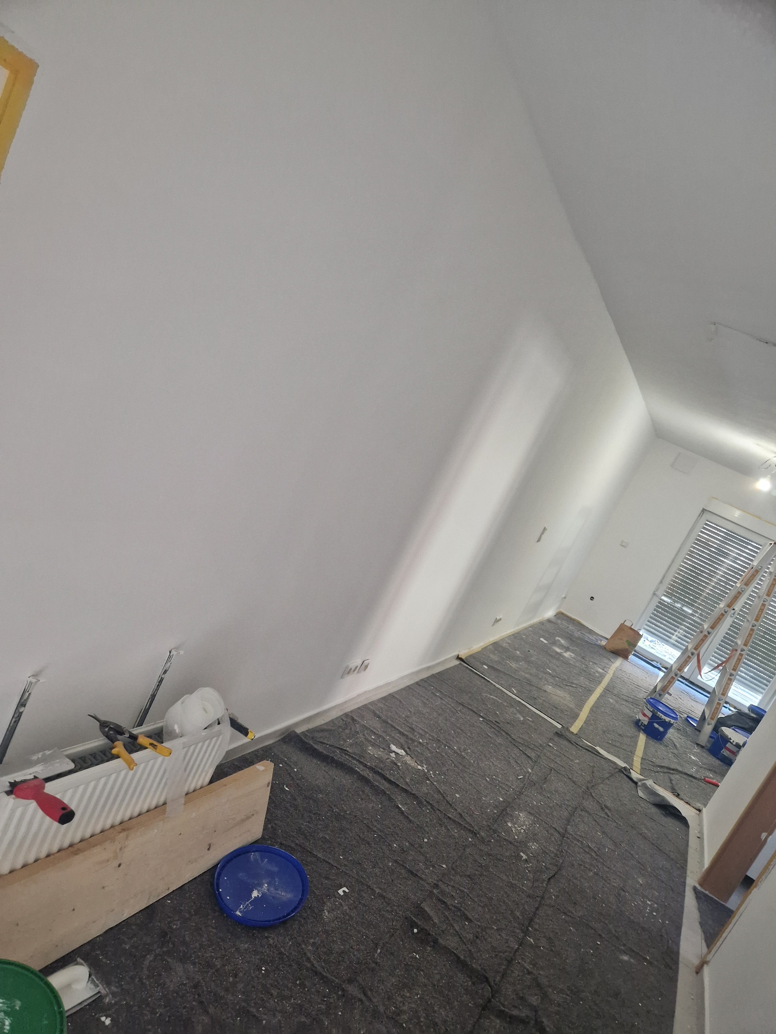 Świeżo pomalowane na biało ściany w pokoju, zabezpieczona podłoga, drabina oparta o okno, widoczne narzędzia malarskie i wiaderka z farbą.