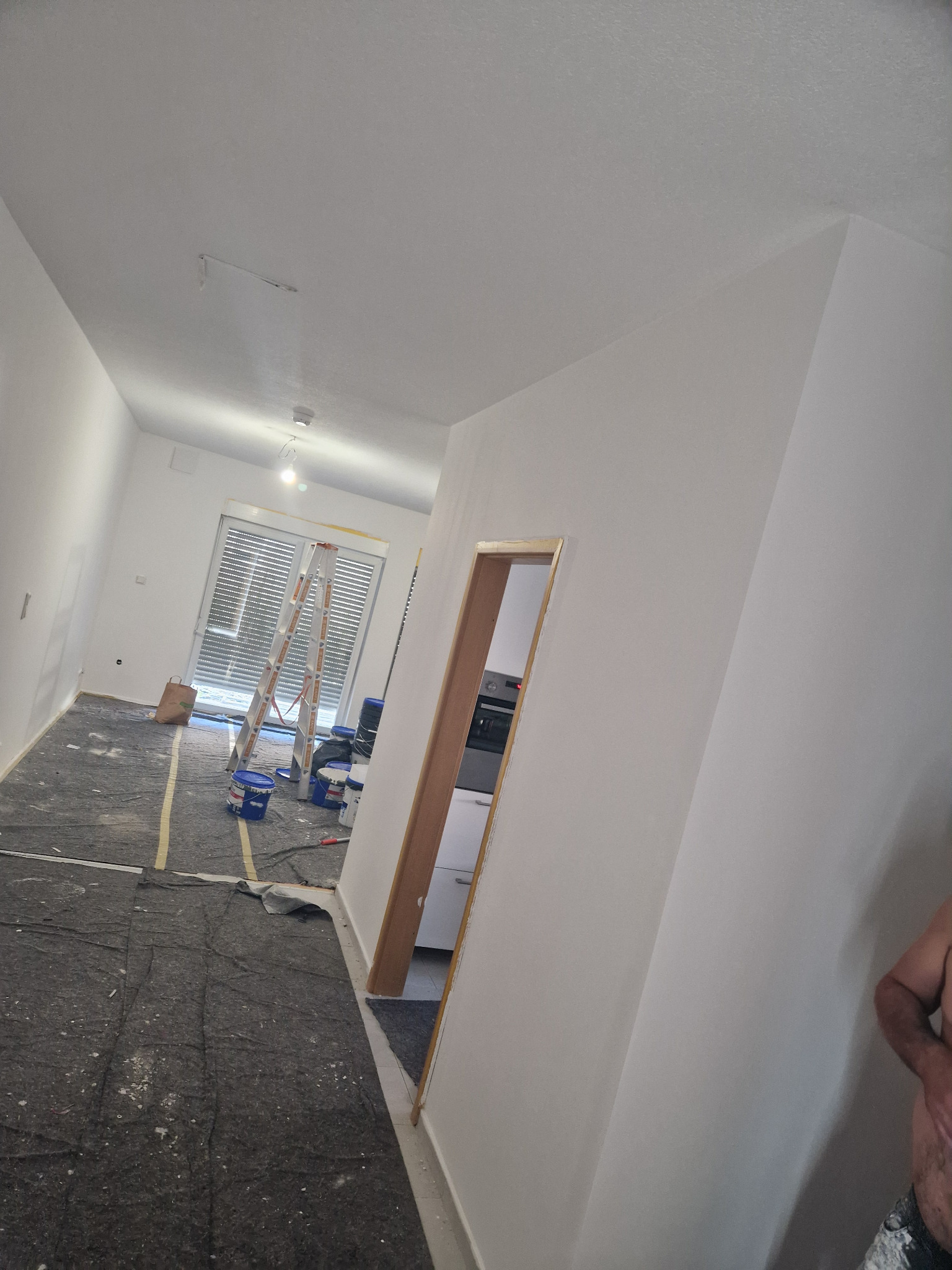 Wnętrze mieszkania w trakcie malowania ścian na biało, widoczna drabina oparta o okno, zakryta podłoga, puszki z farbą i fragment sylwetki malarza.