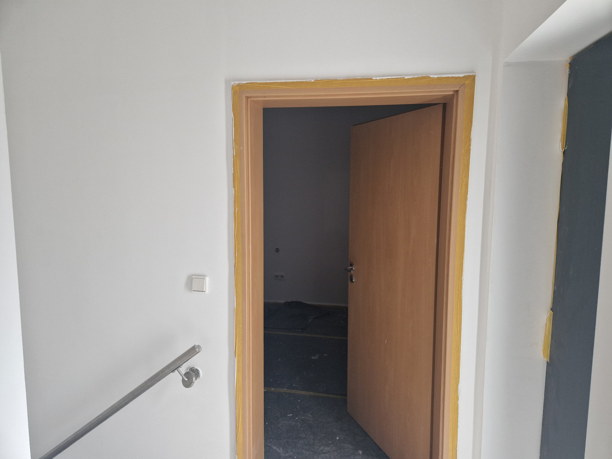 Przygotowanie do malowania ścian i futryny drzwiowej z użyciem taśmy malarskiej w Katowicach. Widoczny fragment schodów z metalową barierką oraz zabezpieczona podłoga w pomieszczeniu za drzwiami.