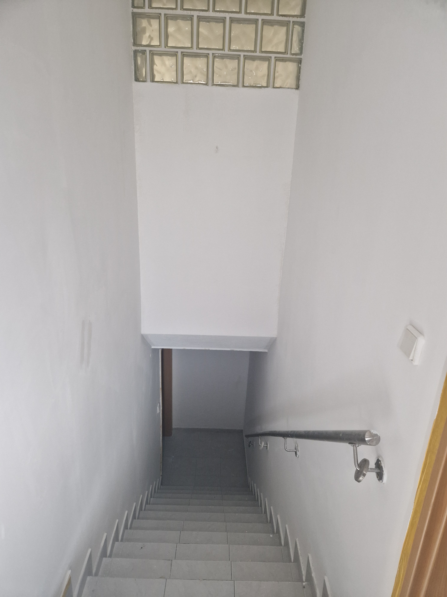 Klatka schodowa z szarymi schodami i metalową poręczą, ściany wykończone na gładko w odcieniach bieli, górna część ściany z luksferami.