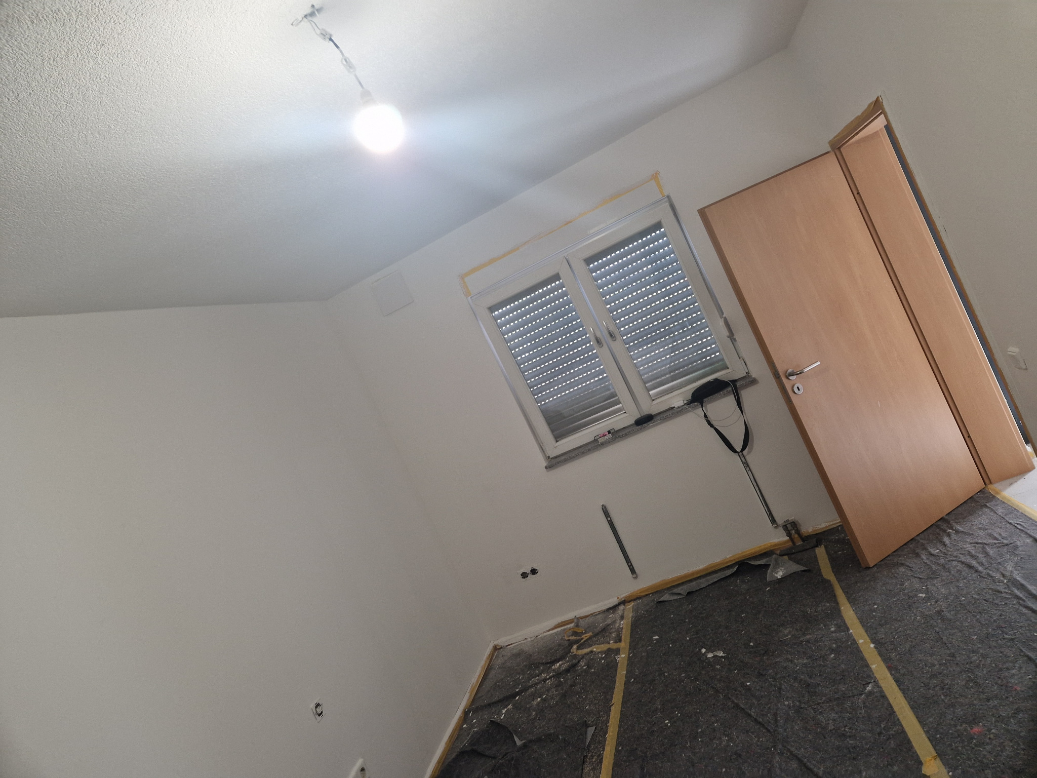 Pokój w trakcie remontu: ściany przygotowane do malowania, okno zabezpieczone taśmą malarską, podłoga zakryta folią ochronną, widoczna wisząca lampa.