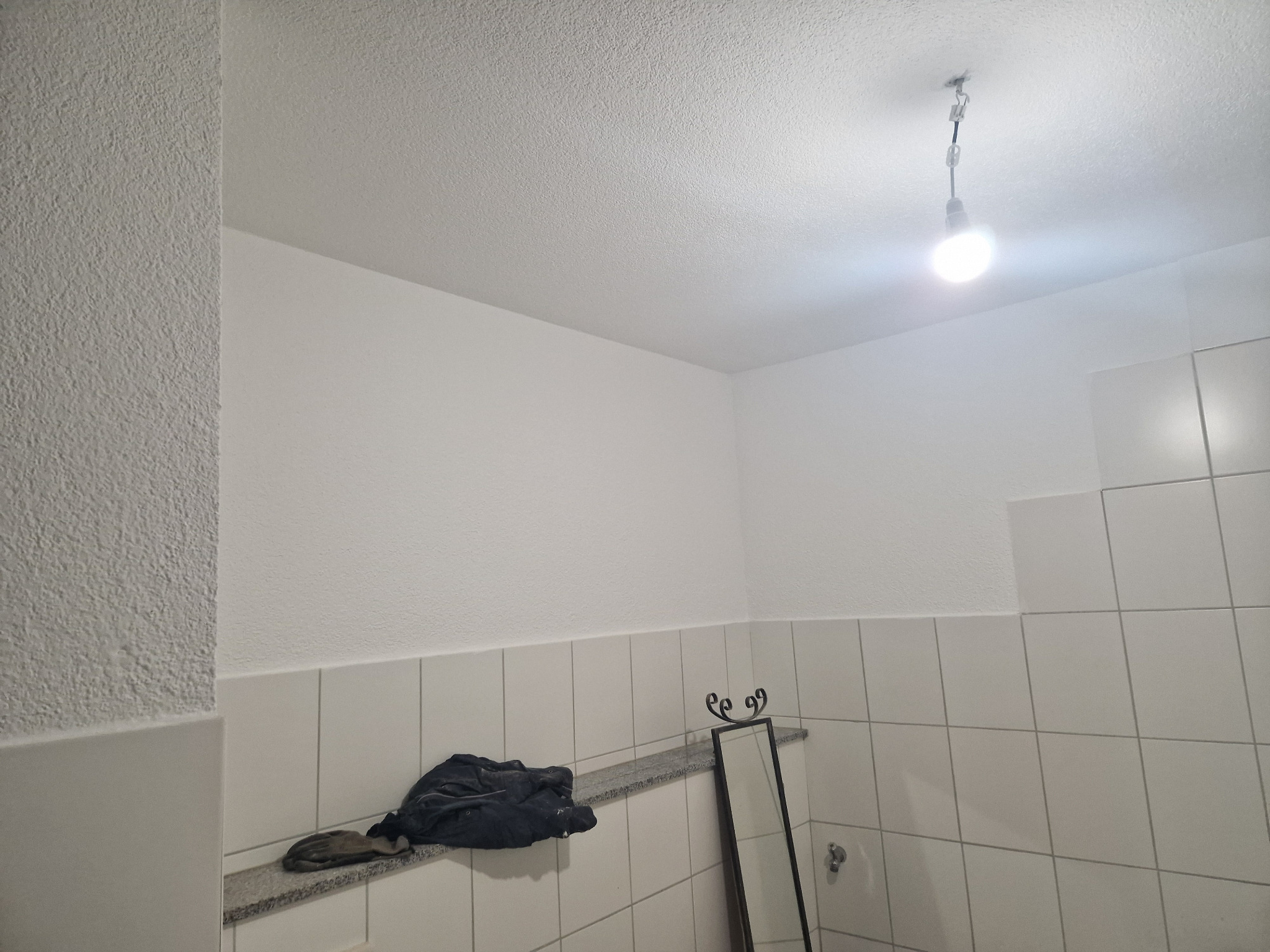 Wnętrze łazienki w trakcie remontu, widoczne białe płytki, surowa żarówka na kablu, oraz pozostałości po pracach budowlanych: ubrudzona odzież robocza i lustro oparty o ścianę.