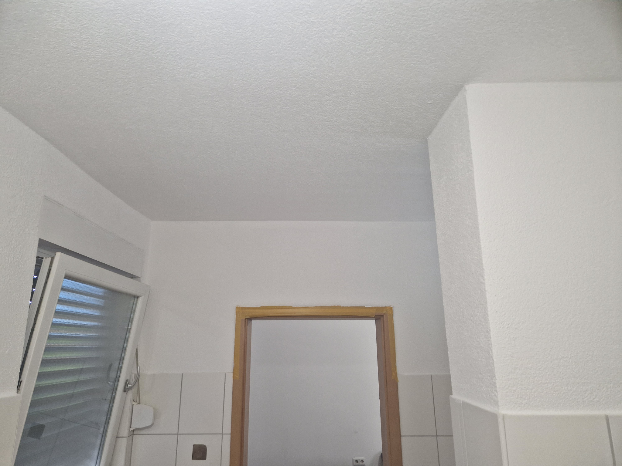 Białe wnętrze po świeżym malowaniu, widoczny sufit z fakturą, fragment okna z żaluzjami i ościeżnica drzwiowa z drewnianym obramowaniem.