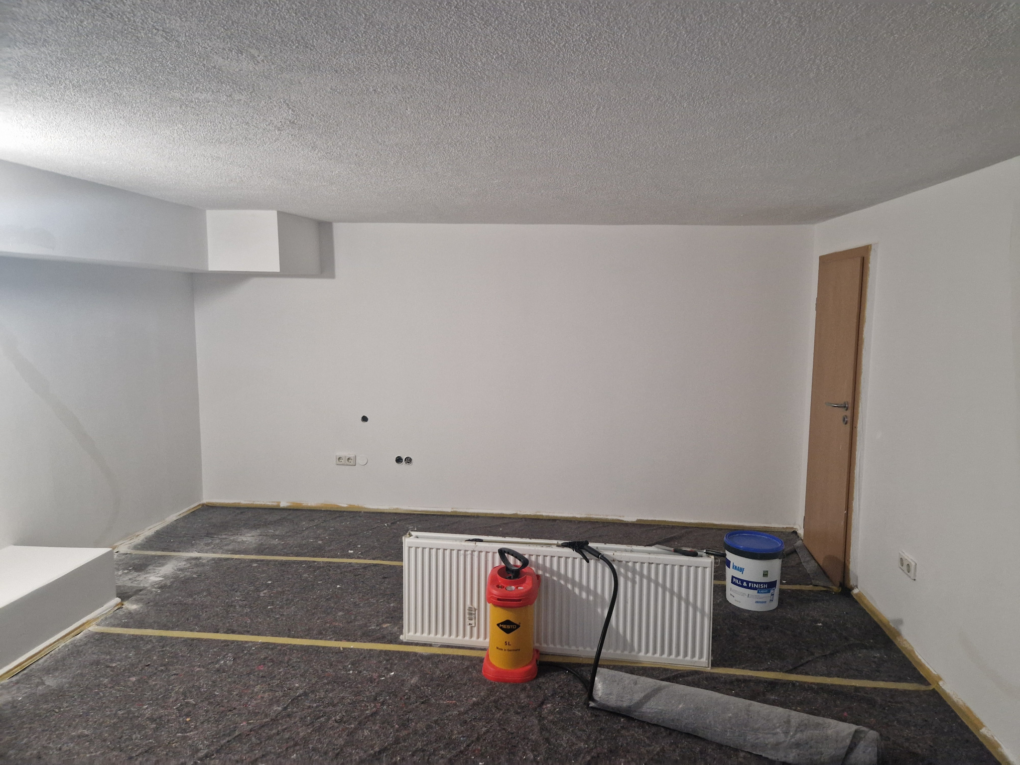 Pokój w trakcie prac wykończeniowych: zabezpieczona podłoga, białe ściany i sufit, grzejnik z opryskiwaczem i wiadro z masą szpachlową.