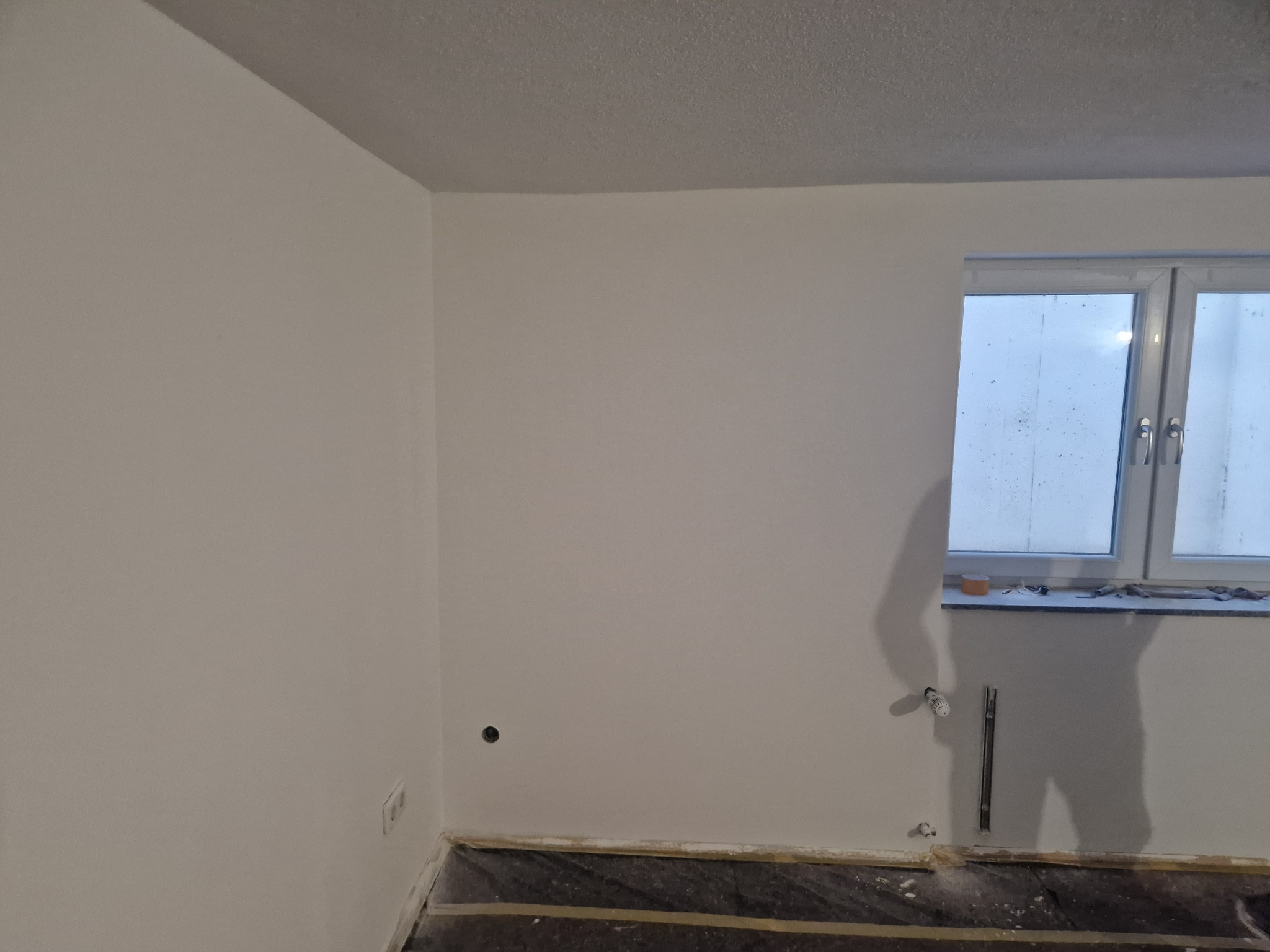 Wnętrze pokoju po malowaniu ścian na biało, widoczne nowe okno PCV, zakryta podłoga i ślady po instalacji grzewczej na ścianie.