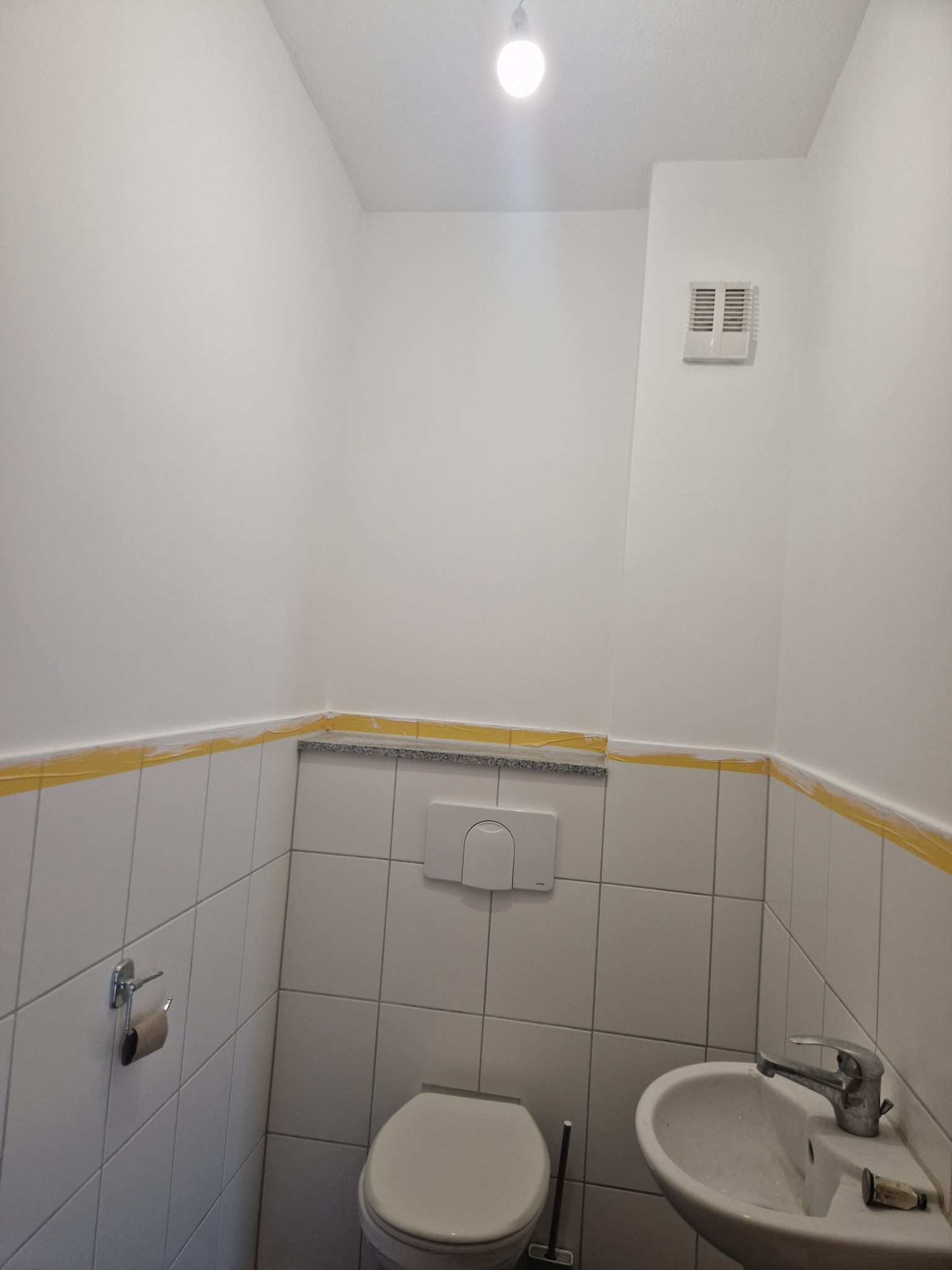 Biała łazienka w trakcie remontu, zabezpieczona żółtą taśmą malarską na styku płytek i ściany, widoczny fragment toalety i umywalki, oraz wentylator na ścianie.