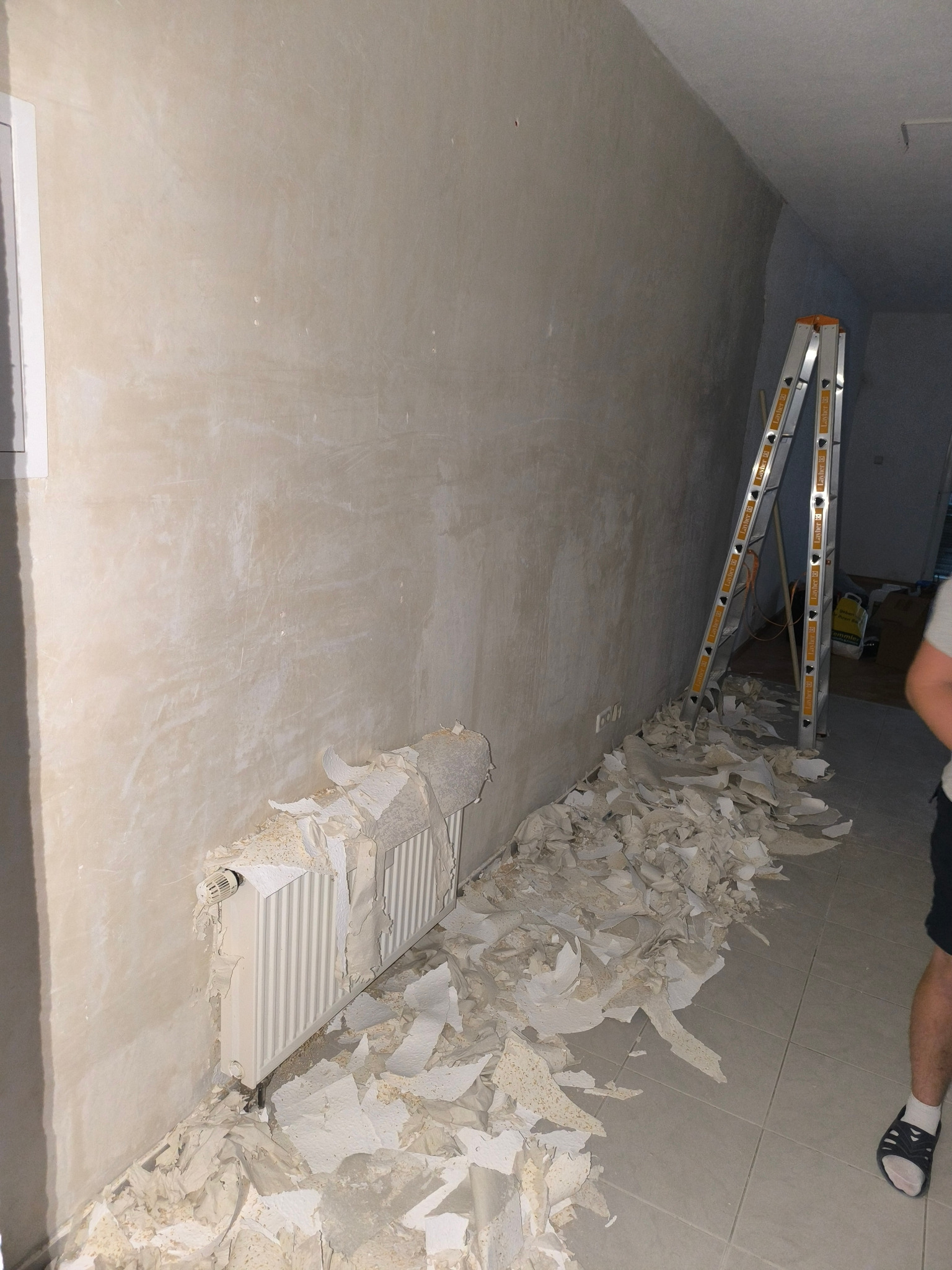 Ściana po usunięciu starej powłoki, z widocznym grzejnikiem częściowo pokrytym resztkami, drabina oparta o ścianę, przygotowanie do dalszych prac wykończeniowych.