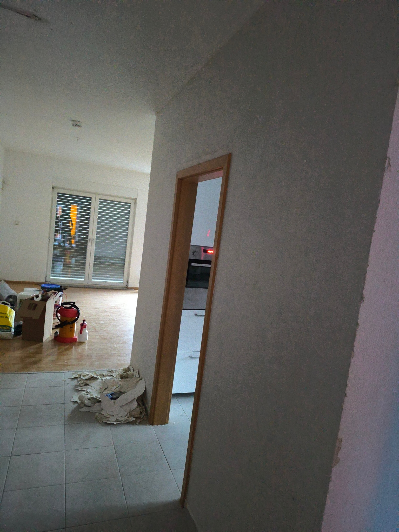 Wnętrze w trakcie remontu: widok z korytarza na pokój z oknem, widoczne narzędzia, worki, oraz fragment kuchni przez otwarte drzwi. Na podłodze w korytarzu leżą resztki materiałów budowlanych.