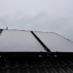 Dwa panele słoneczne, pokryte kroplami wody, zamontowane na czarnym, błyszczącym dachu z dachówki ceramicznej, widoczne elementy montażowe i rura, pochmurne niebo w tle.