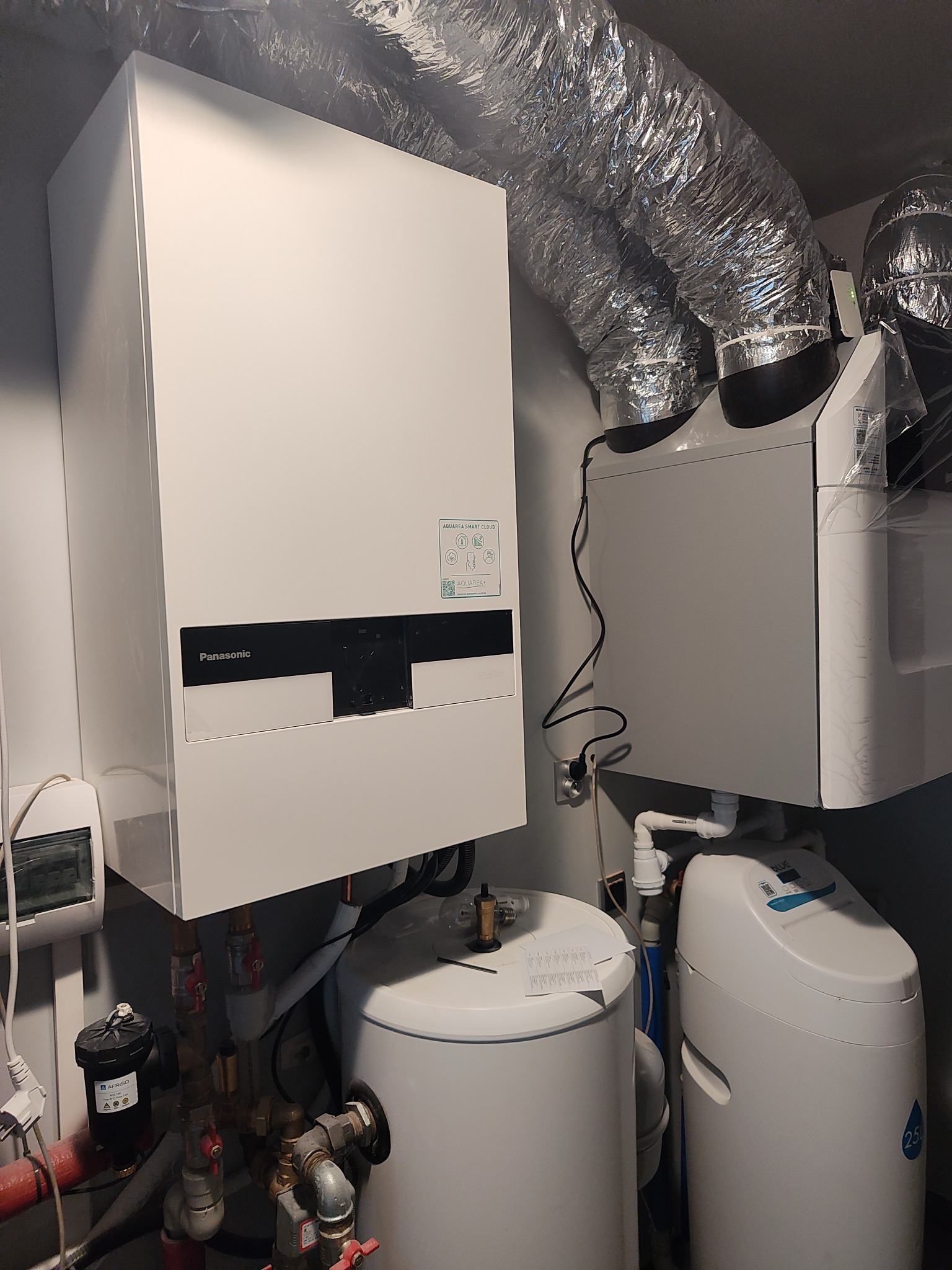 Biały, wiszący piec gazowy Panasonic z podłączonymi rurami i instalacją, widoczne elementy systemu uzdatniania wody oraz wentylacja z folii aluminiowej.