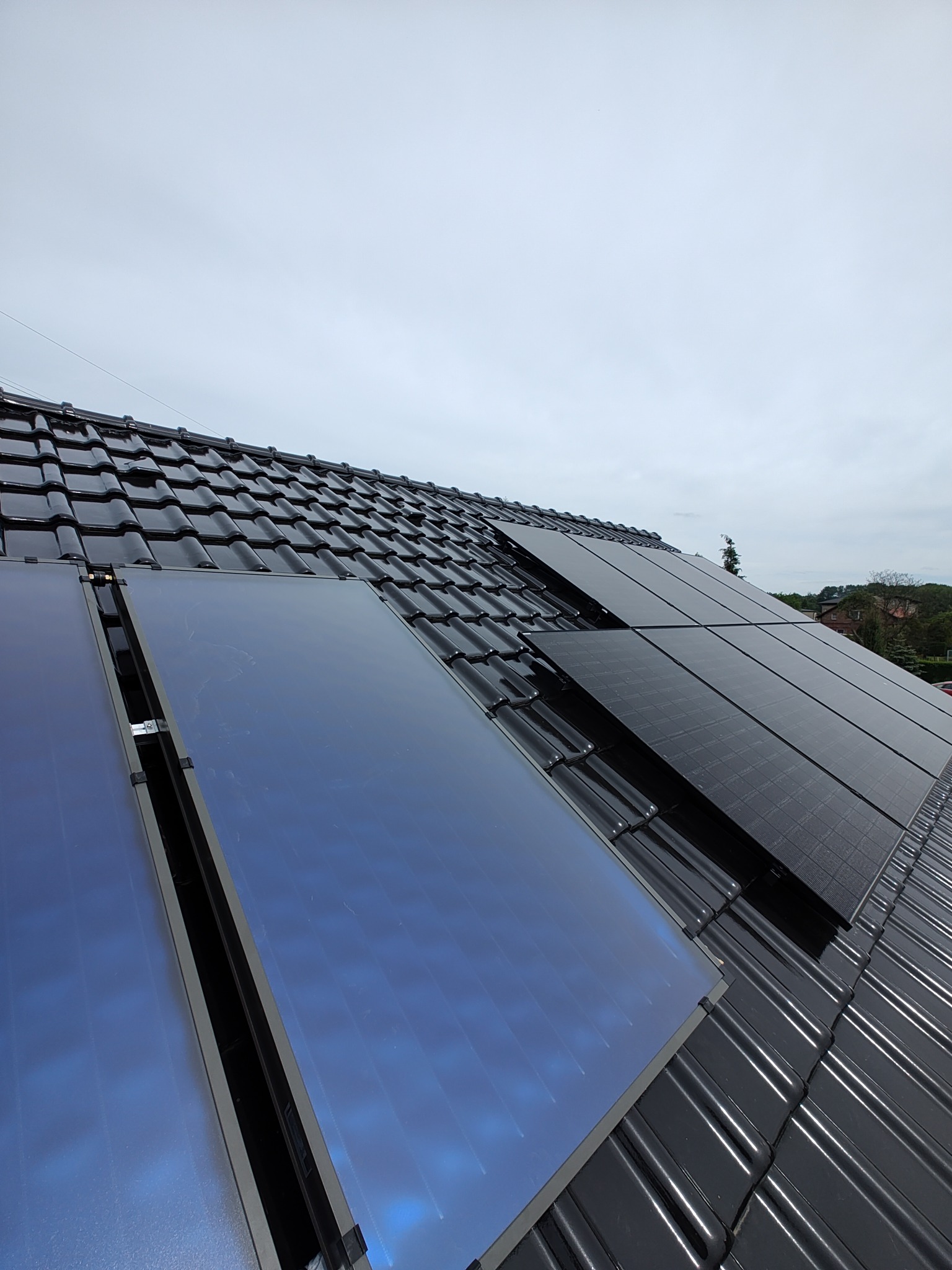 Częściowy widok na dachu z czarnej dachówki, na którym zamontowano niebieskie kolektory słoneczne i czarne panele fotowoltaiczne w pochmurny dzień.