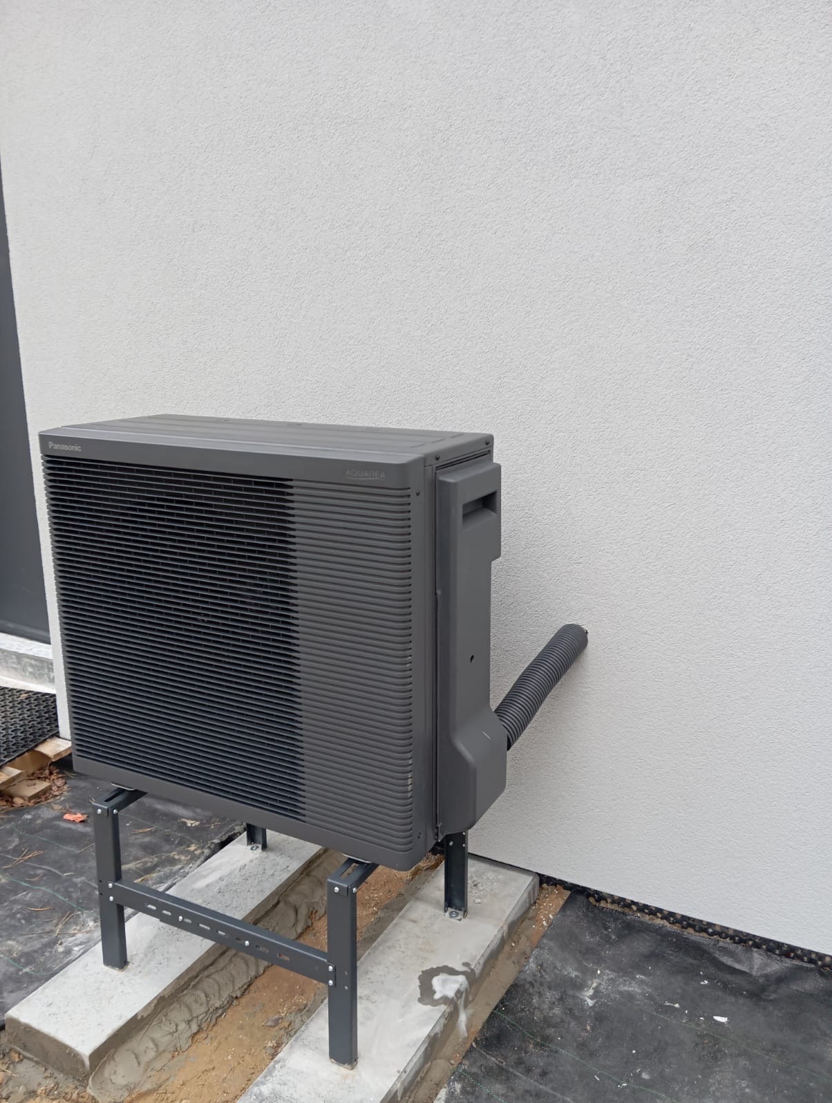 Szara pompa ciepła Panasonic Aquarea zamontowana na metalowym stelażu przy jasnej ścianie budynku, z widoczną rurą odprowadzającą.