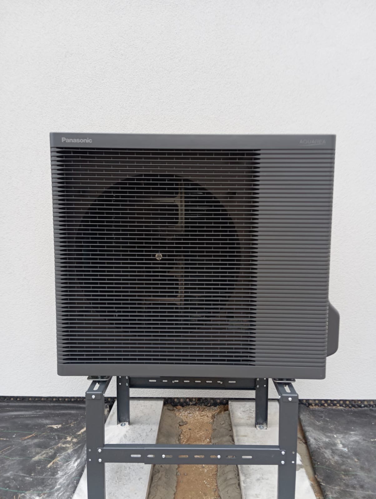 Zewnętrzna jednostka pompy ciepła Panasonic Aquarea, zamontowana na metalowym stelażu na betonowej podstawie w trakcie instalacji.