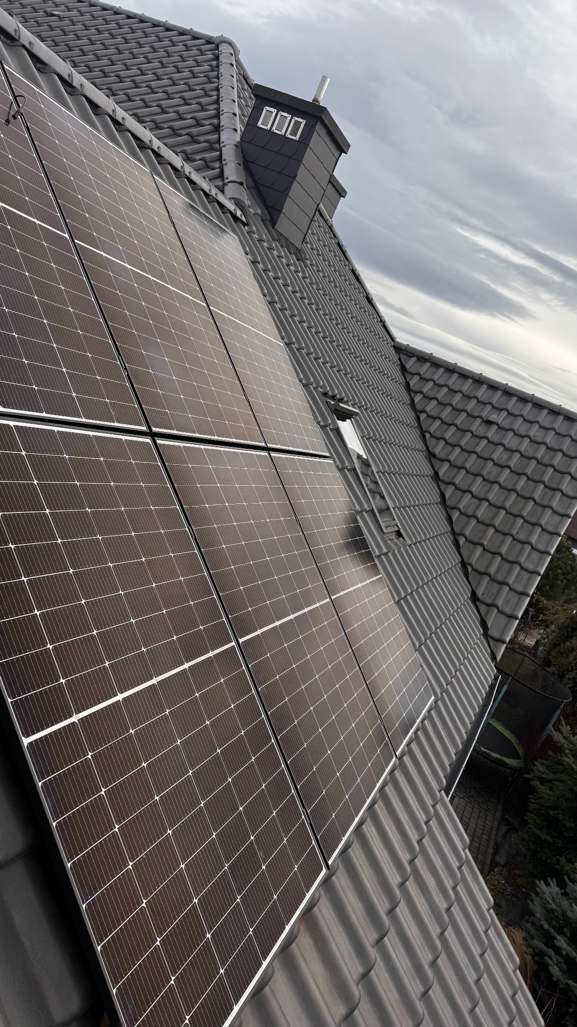 Panele fotowoltaiczne zamontowane na dachu z szarej dachówki ceramicznej, widok z dołu pod kątem, komin w tle.