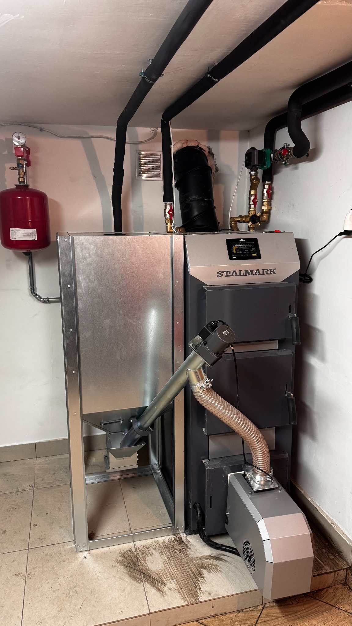 Nowoczesny piec na pellet marki Stalmark z podajnikiem, zamontowany w kotłowni, widoczne rury instalacji grzewczej i ślady po pellet na podłodze.